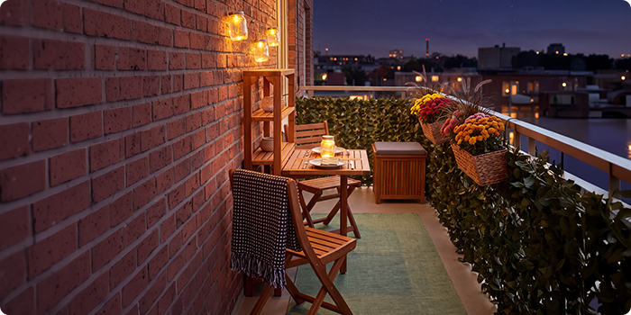Petit balcon avec chaises et table en bois, éclairé par des lanternes et des bougies, décoré de fleurs en pot et de verdure, avec vue sur un panorama urbain de nuit.